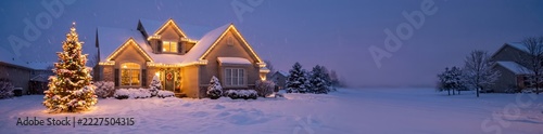 Residential house decorated with glowing warm christmas lights, illuminated pine tree in a snow-covered front yard during a blue twilight winter evening