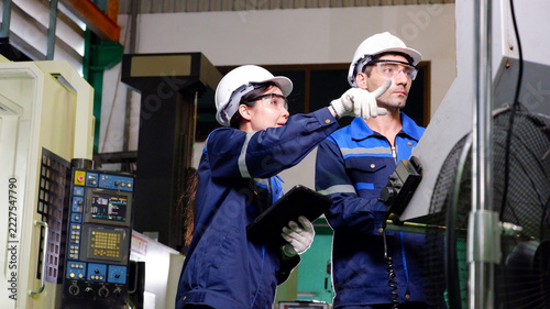 Male and female engineers collaborate to inspect machine components and record operational data inside an industrial factory.