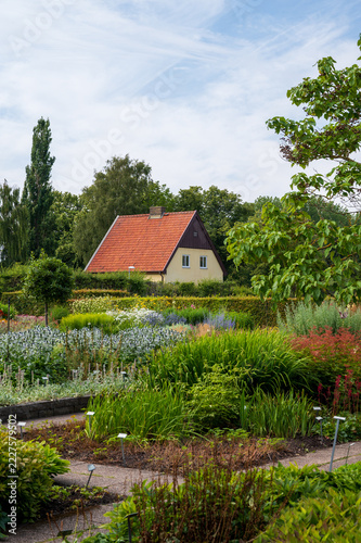 Red roofed house beside a lush perennial garden under a hazy blue sky