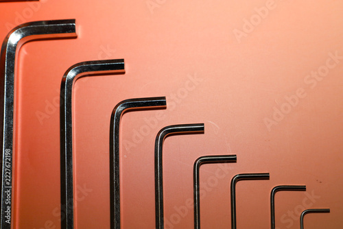 Set of metal hex keys arranged by size on a bright orange and peach gradient background. Chromed tools are laid out in a clear vertical row, creating a concise graphic composition.