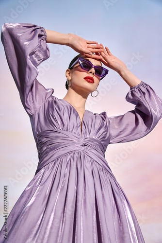 Model poses outdoors wearing a purple dress and sunglasses against a colorful sky during sunset