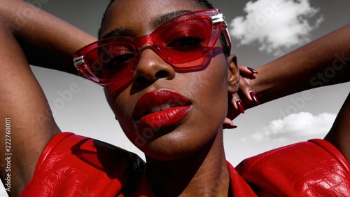 Woman wearing red sunglasses poses against a cloudy sky during daytime while showcasing her bold style