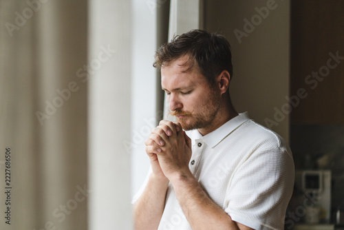 Christian man praying with clasped hands by window