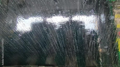 Inside view of car driving through automated car wash machine with water, soap foam and rotating brushes on windshield
