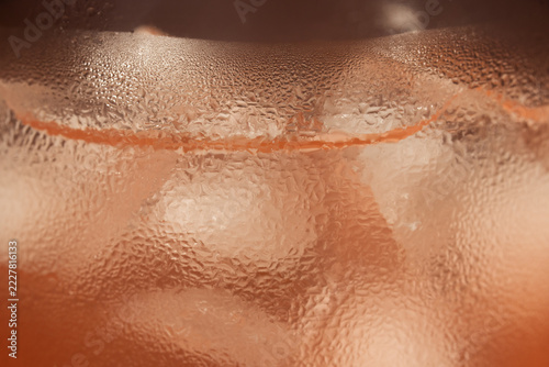 Fotografie Macro close-up of a cold drink glass covered in heavy condensation water droplets, showing ice cubes, warm orange tones, fresh liquid texture, cocktail detail, and summer refreshment concept