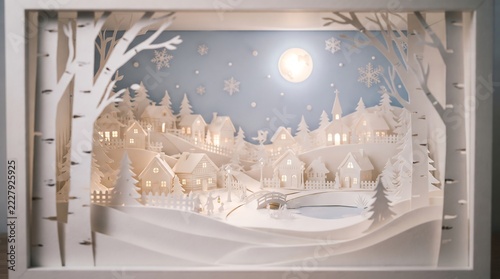 Layered papercut diorama displaying miniature snowy village under glowing full moon during tranquil winter season