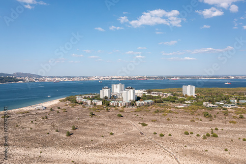 Aerial view of Troia resort towers among sandy dunes in Portugal. A beach lines the blue Sado estuary, with the city of Setubal on the far shore under white clouds.