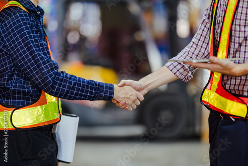 Two workers in reflective safety vests shake hands in a warehouse, symbolizing partnership and agreement in logistics, with industrial equipment blurred in the background.