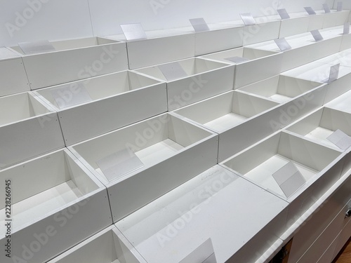 Rows of empty white retail display compartments with transparent label holders arranged diagonally inside a store