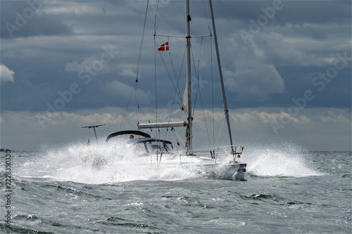 Segelboot kämpft gegen raue Wellen und aufspritzende Gischt auf stürmischer See unter dramatischem, wolkenverhangenem Himmel.