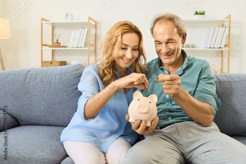 Happy mature couple saving money together. Husband and wife smile on the sofa at home while putting coins into a piggy bank, building family savings for the future. Small steps to big savings.