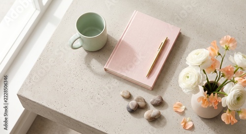 A serene desk setup features a pastel mug, a pink notebook with a pen, and a vase of white and peach flowers. The arrangement creates a calming and elegant workspace atmosphere.
