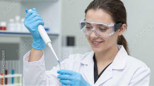 Female laboratory scientist in safety goggles pipetting clear liquid into glass test tube in clinical setting, focused expression and protective gloves