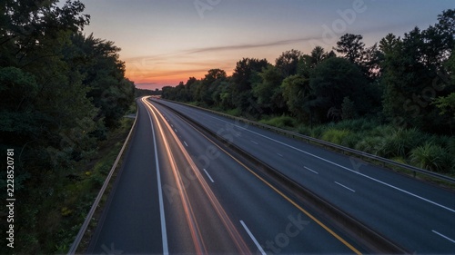 Divided highway at dusk with vehicle light trails and tree-lined verge under pastel sunset sky, road stretching into distance