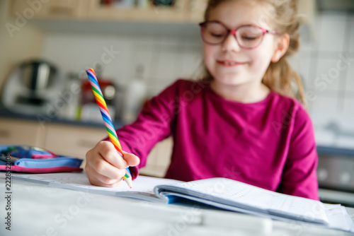 Little school girl learning at home. Elementary class student chid doing maths homework, writing with pencil. Happy pupil with eye glasses