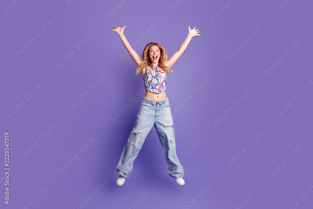 Naklejka premium Happy young woman jumping with arms raised in colorful crop top and wide jeans on a purple background expressing joy and energy