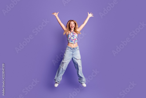Happy young woman jumping with arms raised in colorful crop top and wide jeans on a purple background expressing joy and energy