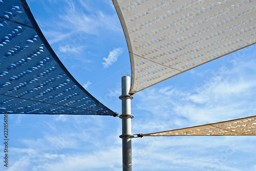 fabric sun shade and awning over patio area. penetrated sun sail spanning above city terrace. blue sky and white clouds. low angle view. metal connector and chain. summer and outdoors concept.