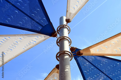 fabric sun shade and awning over patio area. penetrated sun sail spanning above city terrace. blue sky and white clouds. low angle view. metal connector and chain. summer and outdoors concept.