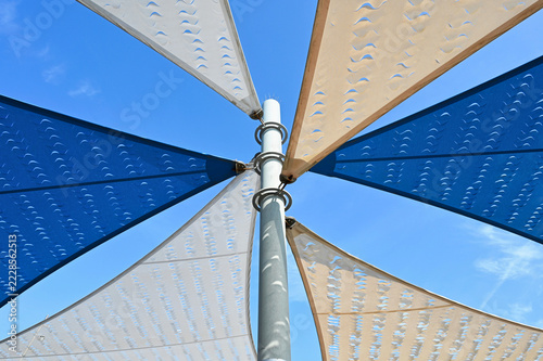 fabric sun shade and awning over patio area. penetrated sun sail spanning above city terrace. blue sky and white clouds. low angle view. metal connector and chain. summer and outdoors concept.