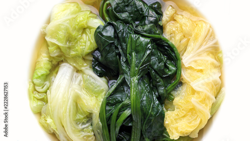 overhead, vibrant culinary shot captures three varieties of boiled leafy green vegetables arranged neatly in sections inside round bowl, submerged slightly in clear, light broth, white background