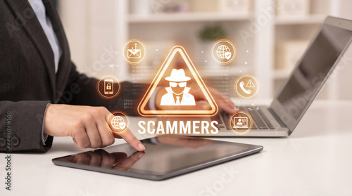Business professional using digital tablet with glowing scammers warning hologram and cybersecurity icons illustrating online fraud and phishing threats.