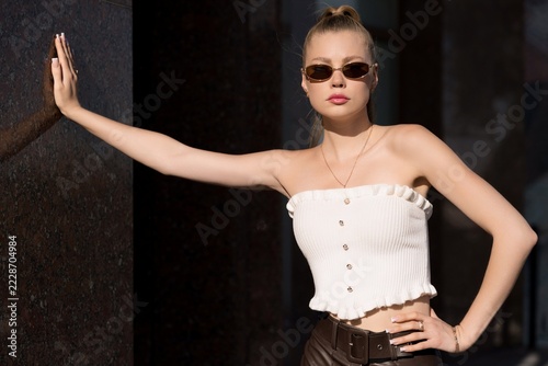 Half body portrait of a young caucasian blonde woman posing against a stone wall on a sunny day, wearing white top, leather pants, and sunglasses.
