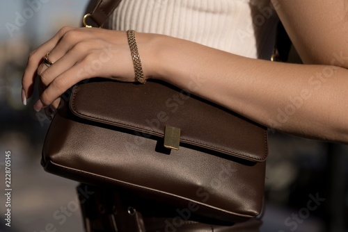 Close-up fashion shot of a young caucasian woman wearing white off-shoulder top and leather pants, holding a brown handbag on a sunny day outdoors.