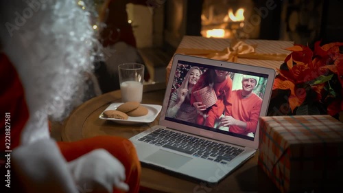 Santa Claus video calling family with little girl on laptop, greeting child by webcam, opening Christmas gift box in virtual online meeting using computer sitting in his decoration residence with