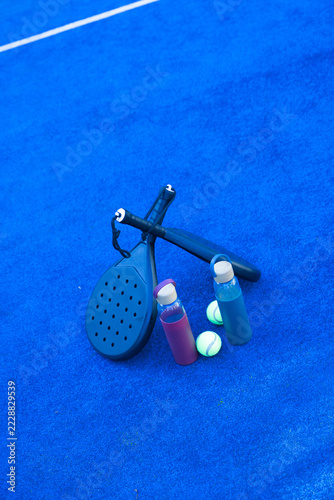 Two black paddle rackets are resting crossed on blue court with balls and bottles, copy space