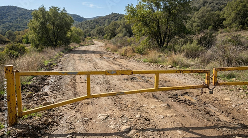 Yellow metal gate blocks dusty rural dirt road overgrown hillside dry vegetation remote landscape no entry