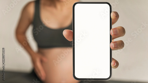 Fotografie Expecting Mother Holding Smartphone With Blank Screen In Front Of Pregnant Belly, Standing In Cozy Home Environment