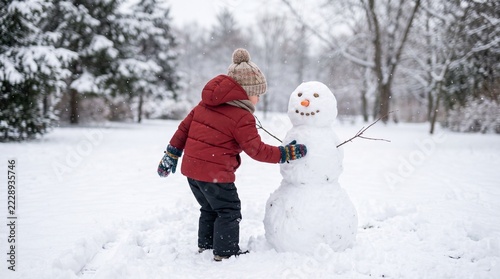 Young child in warm winter clothing building a snowman in a snowy park during snowfall