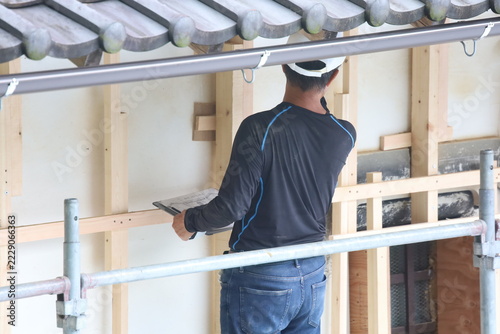 Construction Worker in White Helmet Holding Trowel on Scaffolding with New Drywall and Traditional Tile Roof