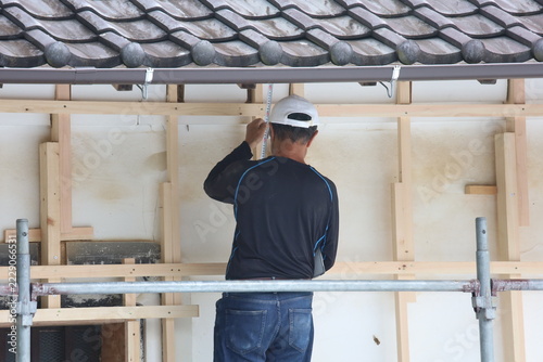 Construction Worker Using Tape Measure on New Wall from Scaffolding Under Traditional Japanese Curved Tile Roof
