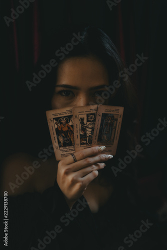 Fotografie Asian fortune teller holding three tarot cards covering her face, mysterious gaze in dark gothic background