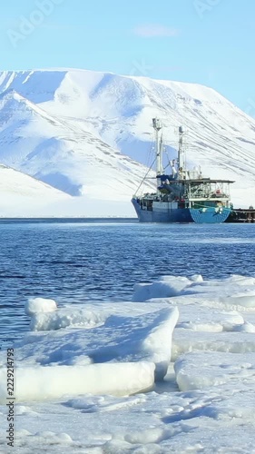 A ship anchored in the blue waters of the Arctic Ocean near sea ice with snow-covered mountains in the background in Svalbard Norway