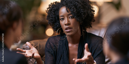 A woman with curly dark hair, wearing a black top, gestures with her hands while engaged in a conversation with two other people, suggesting an intense discussion or meeting