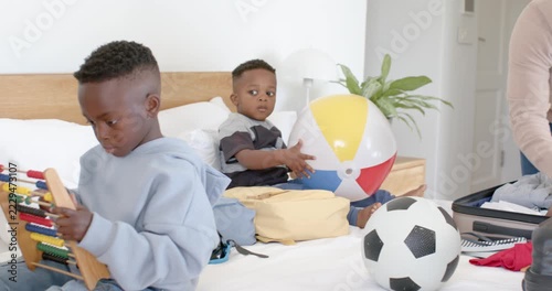 African American family dad packing suitcase for trip on bed while sons tossing ball, using abacus