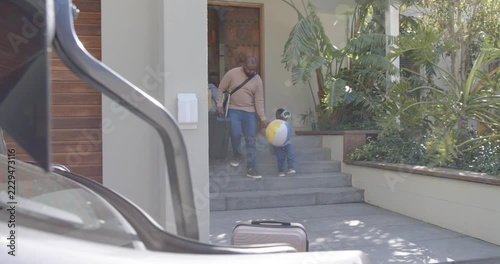 African American father pulling suitcase, guiding son holding beach ball from steps to open trunk