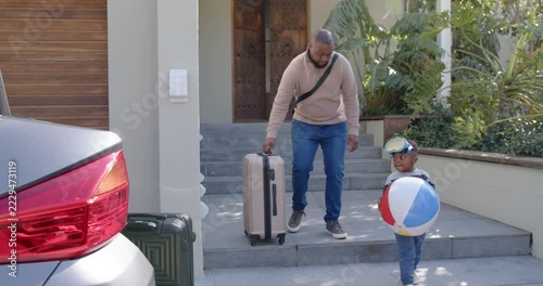 African American dad pulling suitcase at steps while child holding beach ball and goggles for beach