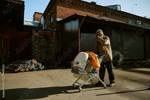 Fotografie Homeless senior Caucasian man pushing shopping cart with belongings along urban street
