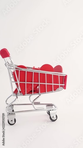 A hand pushes a red miniature metal shopping cart filled with red heart shapes on white background. Cart enters from right, moves left, stops centered, love and Valentine concept.