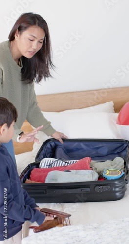 Vertical video: Asian mother and child packing suitcase on bed for trip, zipping, toy plane left