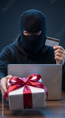 Masked cybercriminal holding credit card behind laptop and wrapped christmas gift representing online shopping fraud.