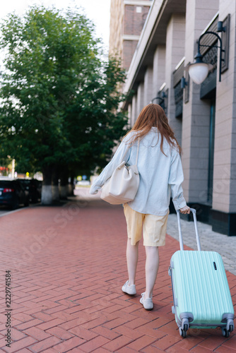 Back view of woman with red hair walking on brick sidewalk, redhead female pulls mint green suitcase and carries handbag. Urban setting suggests travel, vacation, relocation. Natural daylight.