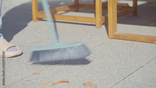 Close up of broom sweeping dry fallen leaves from patio tiles. Brush moving across paved terrace near wooden outdoor furniture. Patio cleaning