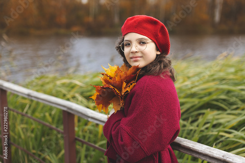 Young girl wearing red knitted beret, glasses, and burgundy coat stands on wooden bridge or pier, holding bouquet of yellow and orange autumn maple leaves against blurry river background