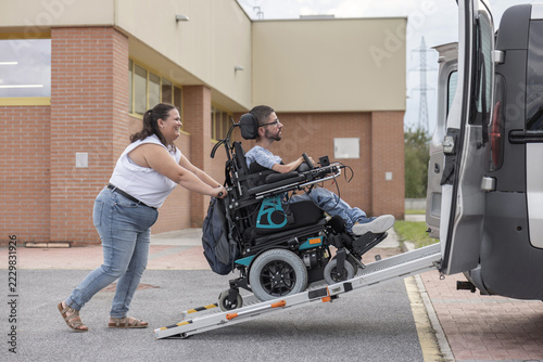 At a building entrance, a woman assists a man in a wheelchair as he boards an adapted van on a ramp during the day.