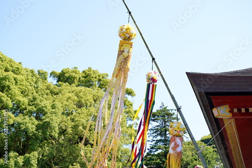 Obraz na plátně 晴天の青空の下、神社などの境内にある竹竿に色鮮やかな七夕飾りの吹き流しと花のボールが吊るされ、緑豊かな木々と日本の伝統的な建物の屋根が調和した夏の風景。